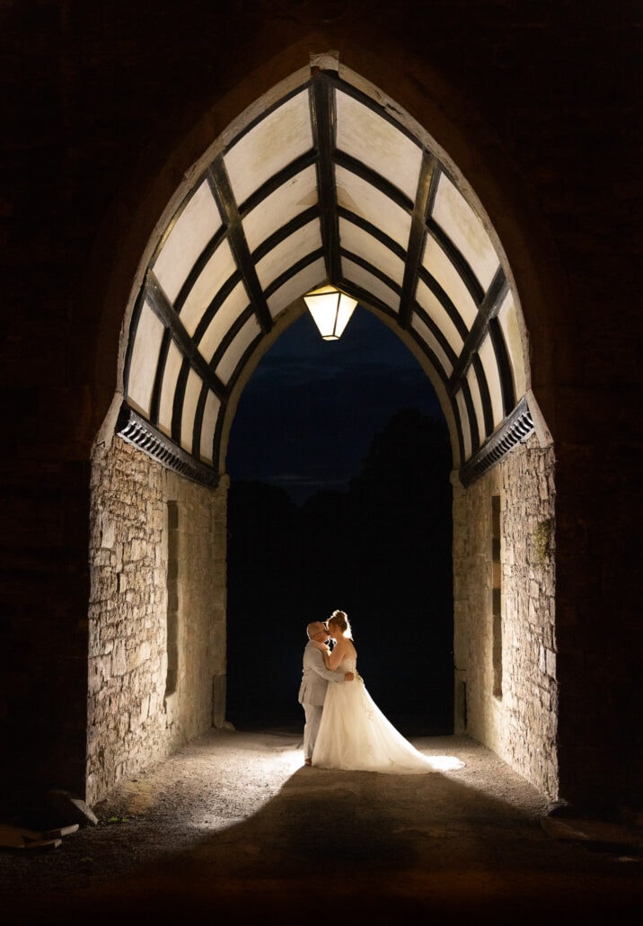 A south wales wedding photographer captures a bride and groom kissing under an archway at night.