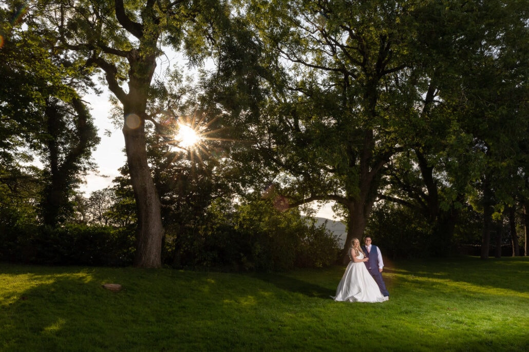 A south wales wedding photographer captures a bride and groom standing in a field with trees in the background.