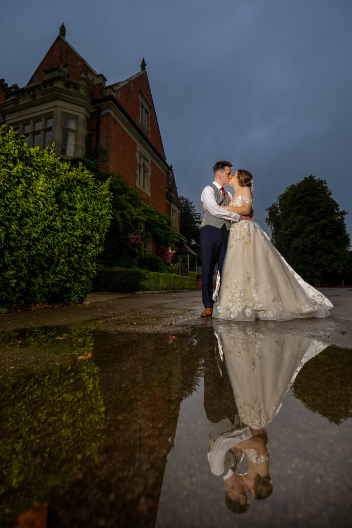 A beautiful bride and groom sharing a romantic kiss in a puddle during their South Wales wedding, with the stunning backdrop of an elegant mansion.