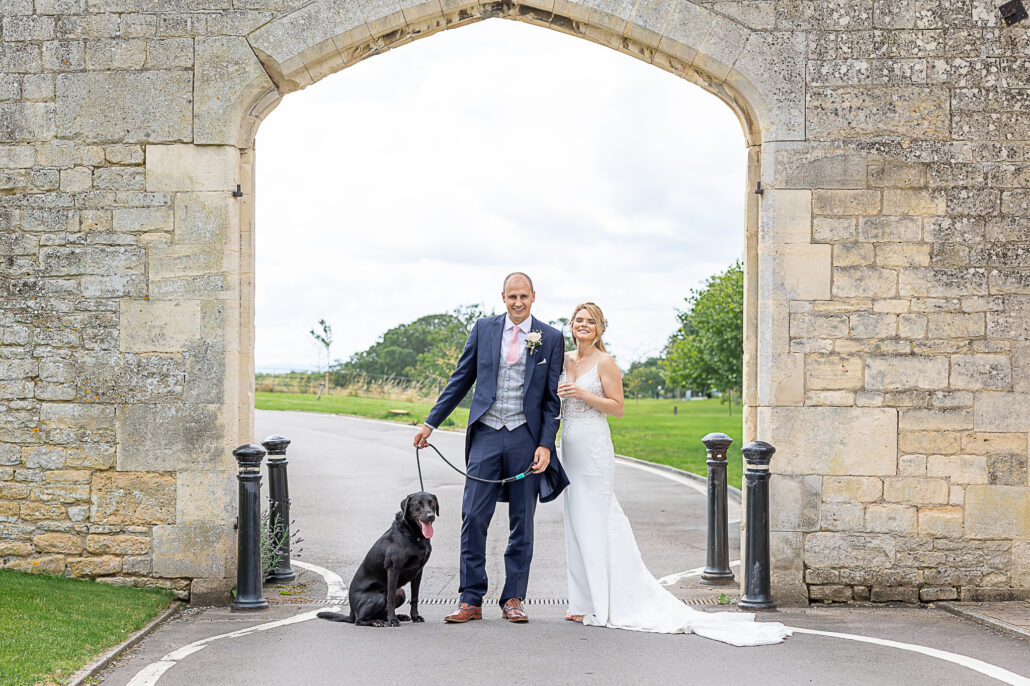 A bride and groom standing in front of an archway with a dog.