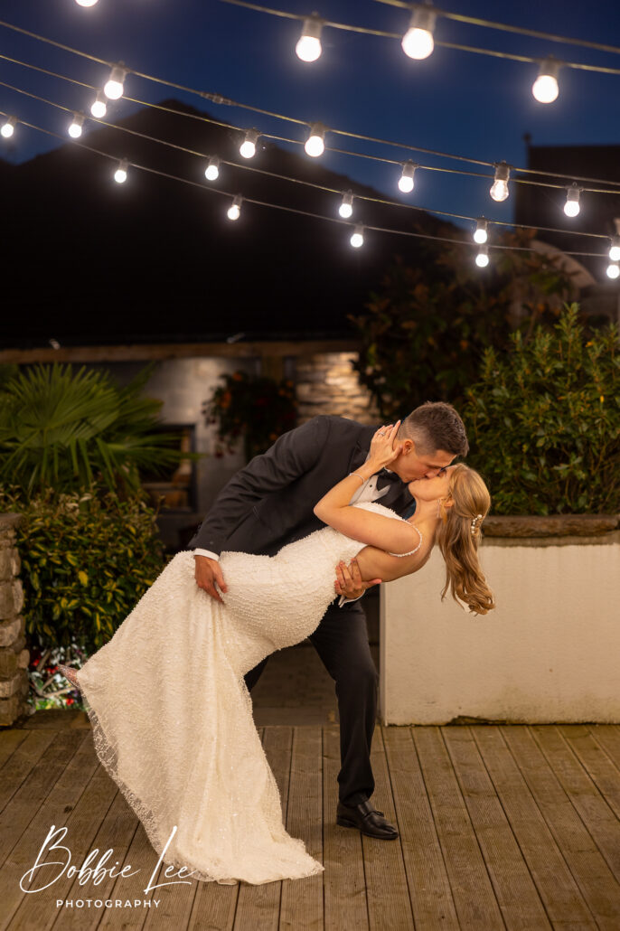 A bride and groom kissing under string lights on a deck.
