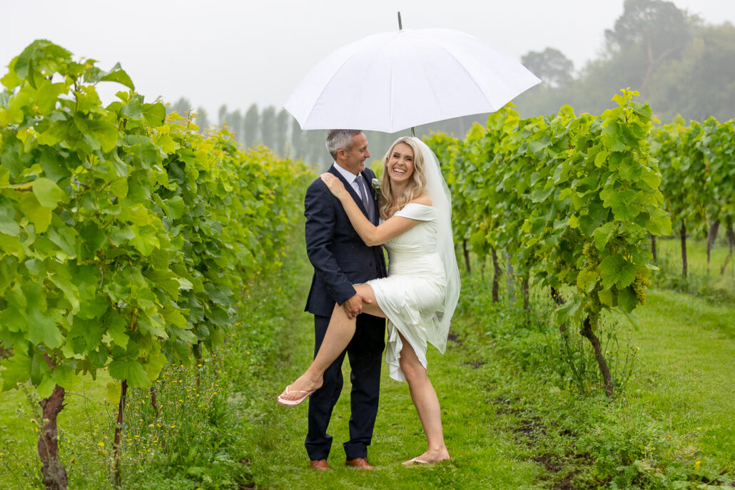 A bride and groom kiss under an umbrella in a vineyard.