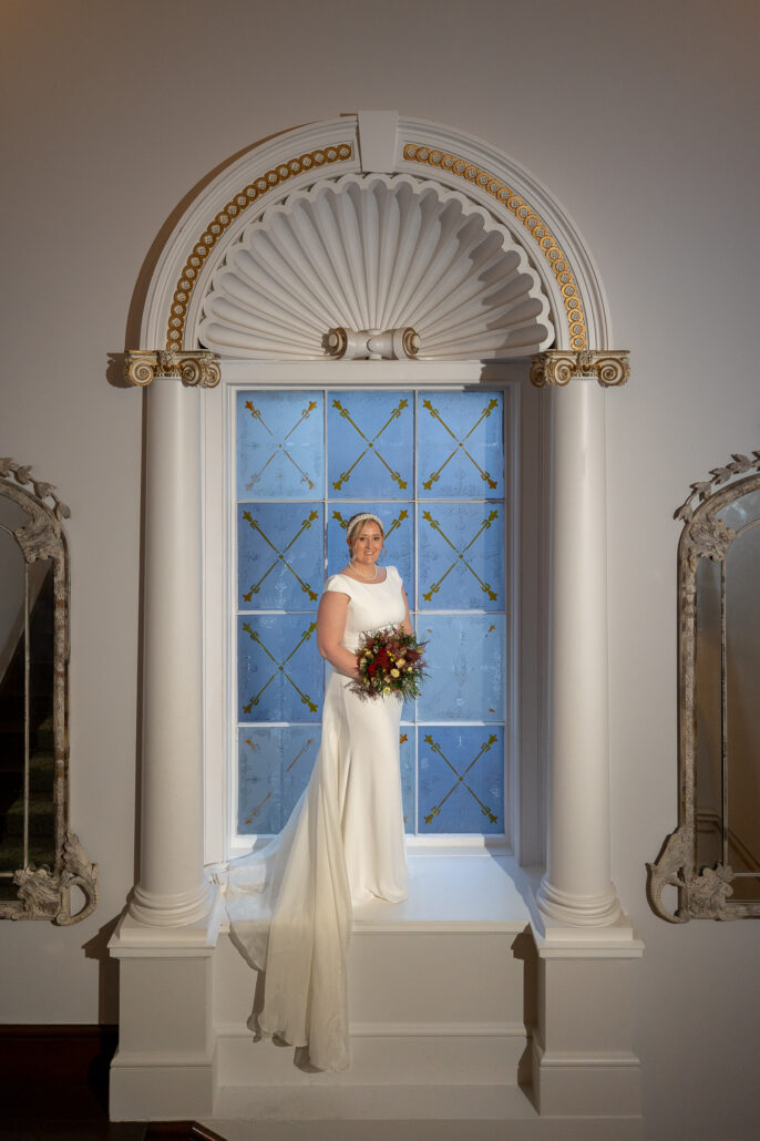 A bride in a white dress standing in front of a window.