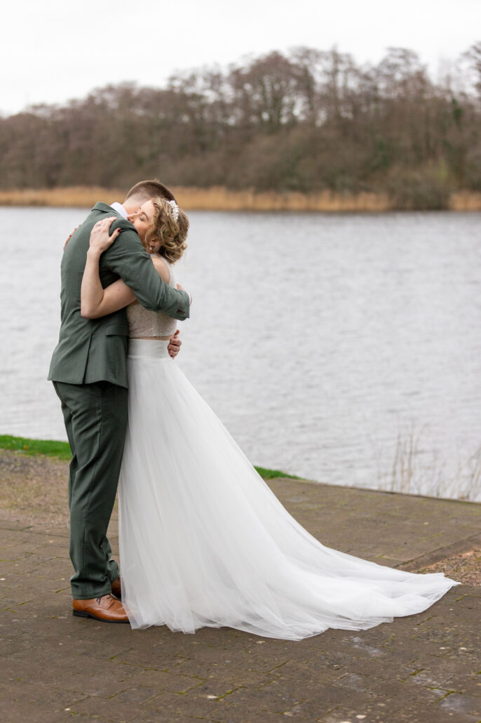 A bride and groom hugging in front of a lake.