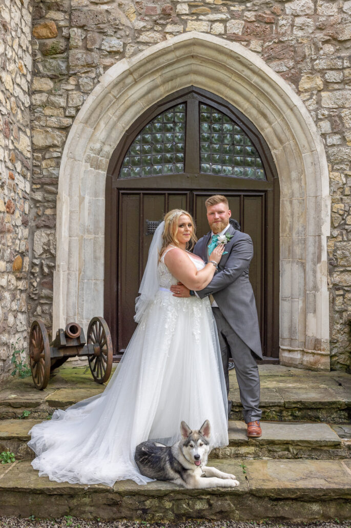 A bride and groom posing with a husky in front of a historic building's arched doorway.