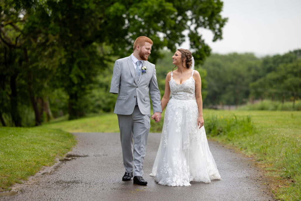 A couple dressed in a wedding gown and suit walk hand in hand on a path in a lush park, with trees and grass in the background, beautifully captured by a South Wales wedding photographer.