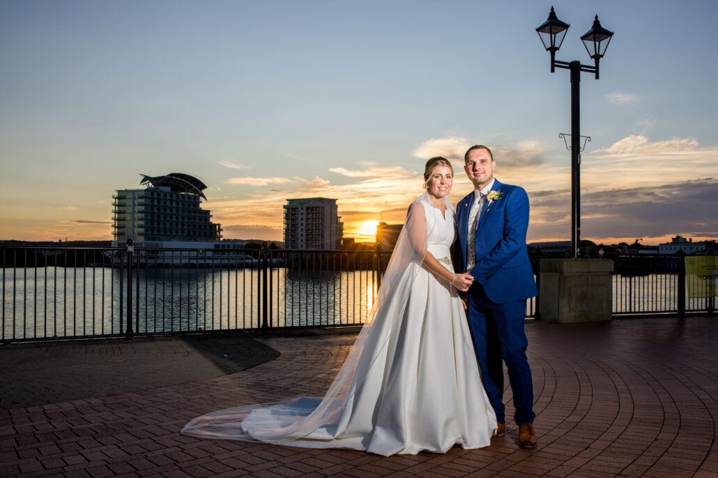 A bride and groom stand hand in hand on a waterfront promenade at sunset, beautifully captured by a South Wales wedding photographer, against the backdrop of modern buildings and an elegant lamppost.