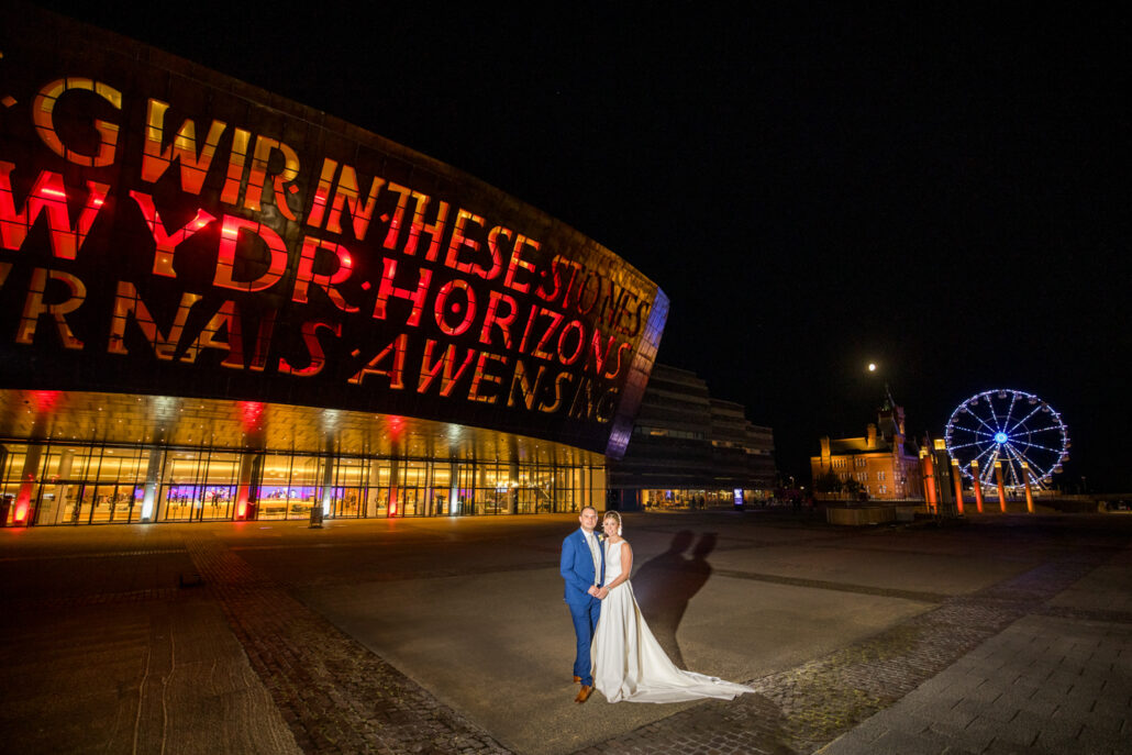A bride and groom stand in front of a large building illuminated with red and yellow text at night, captured perfectly by a South Wales wedding photographer, with a Ferris wheel and another illuminated structure visible in the background.