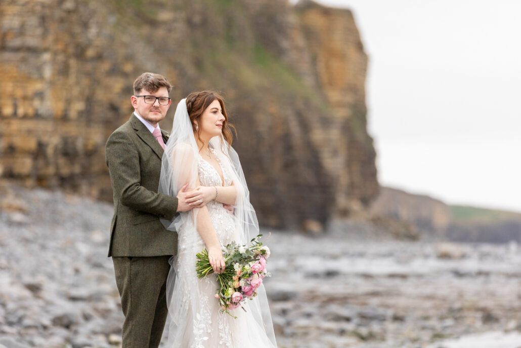 A newlywed couple stands on a rocky beach, with the groom in a suit and the bride in a wedding dress holding a bouquet. Large cliffs are visible in the background, captured beautifully by a South Wales wedding photographer.