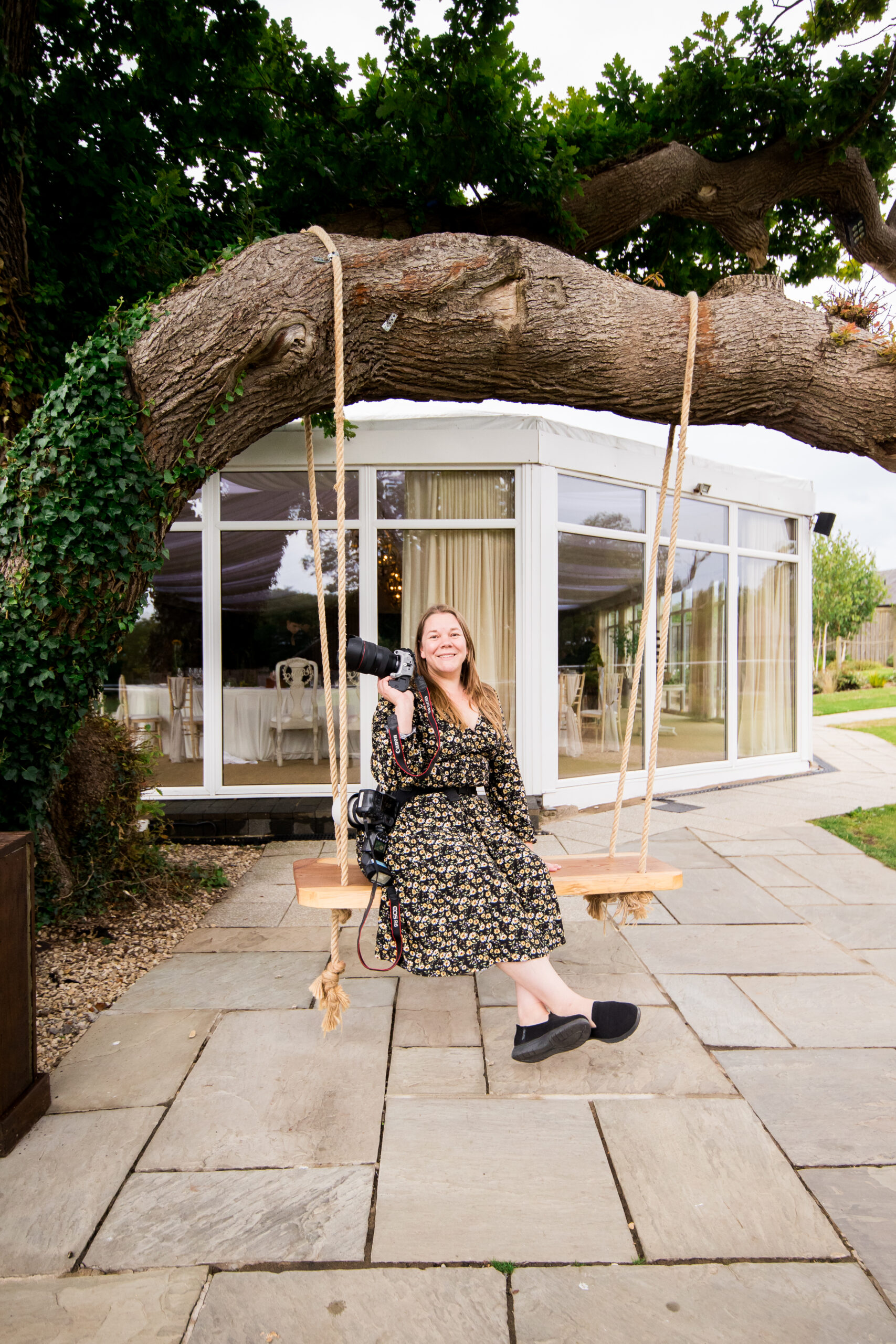 A person sits on a tree swing holding a camera, smiling. They are in front of a glass-walled building surrounded by trees and stone pavement.