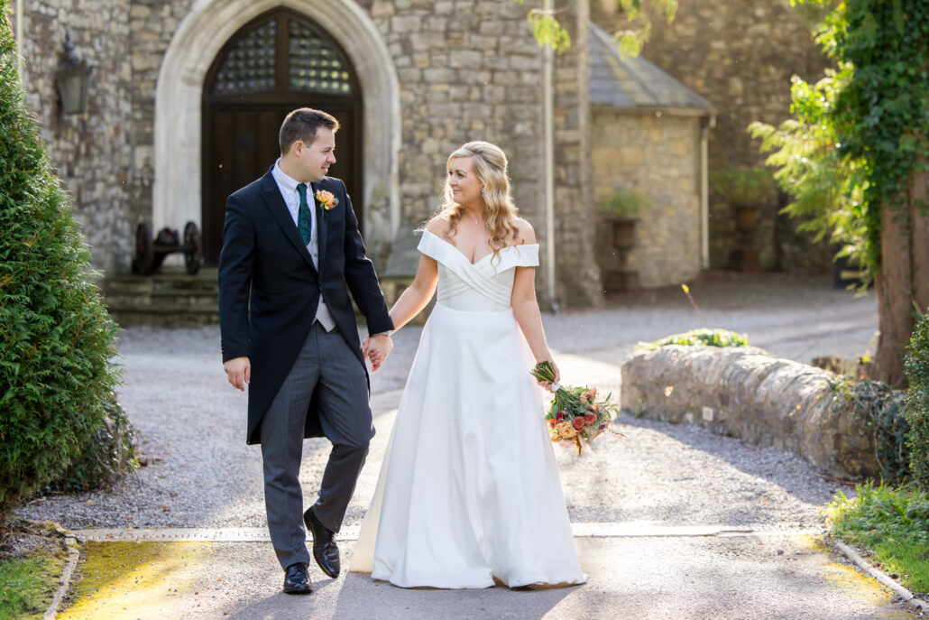 A bride and groom hold hands while walking on a sunny path outside a stone building, beautifully captured by a South Wales wedding photographer. The bride holds a bouquet.