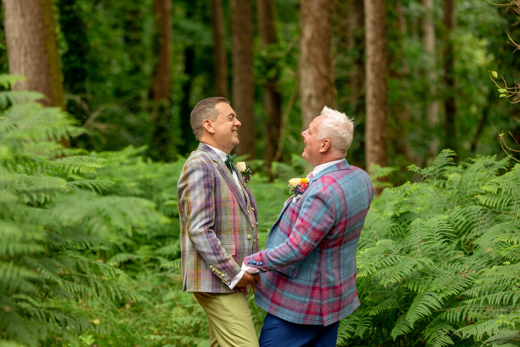 Two people in plaid jackets hold hands and smile at each other, captured by a talented South Wales wedding photographer, standing amid lush greenery in a forest setting.