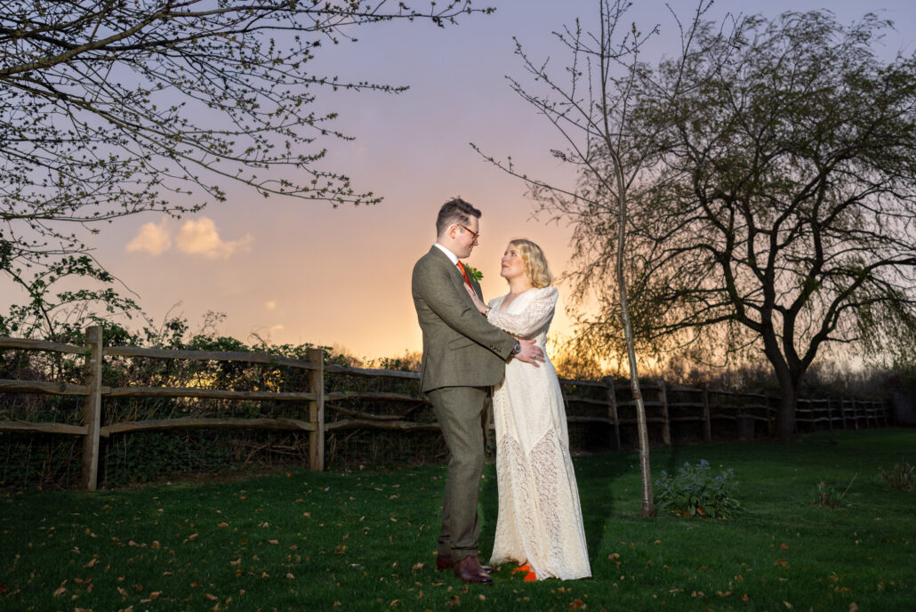 A couple in formal attire stands on grass near a tree, holding each other closely as the South Wales sky paints a sunset of orange and purple hues. Captured by a wedding photographer, a wooden fence lines the scene's edge.