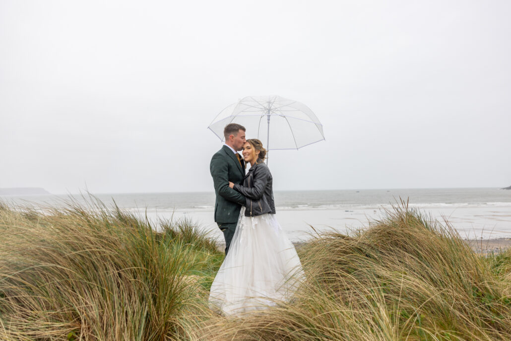 A couple stands under a transparent umbrella in tall grass, framed by the cloudy beach backdrop. Captured with the artistry of a South Wales wedding photographer, this scene tells a story of love and nature intertwined.