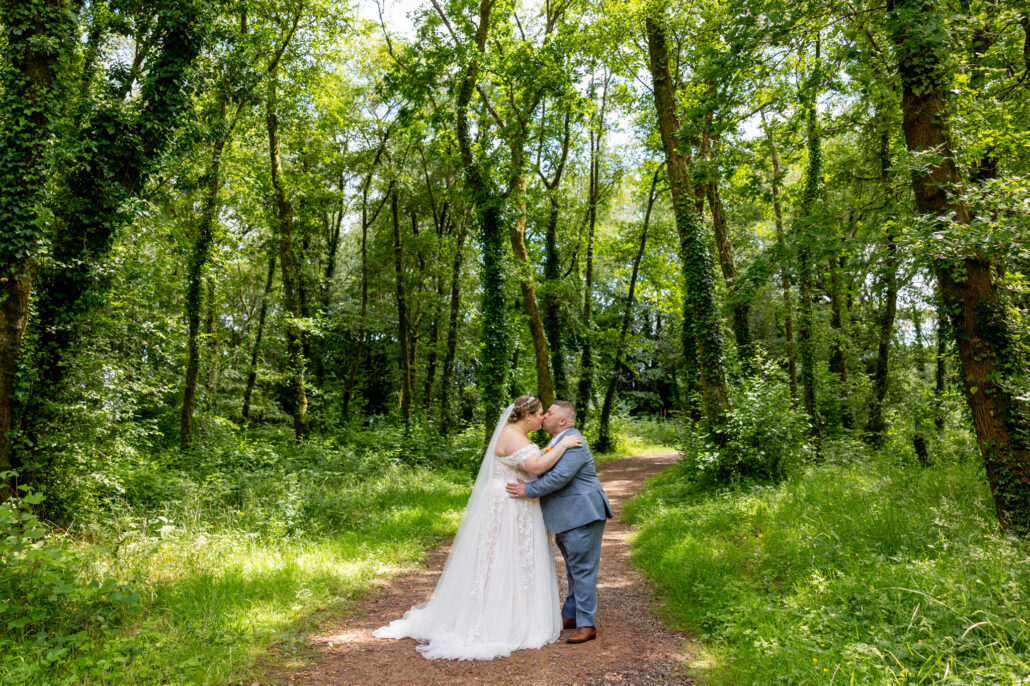 A South Wales wedding photographer captures a bride and groom sharing a kiss on a wooded path, enveloped by lush greenery.