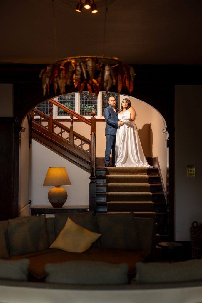 A couple in wedding attire stands on a staircase in a dimly lit room, captured beautifully by a South Wales wedding photographer, with a lamp and a round couch in the foreground.