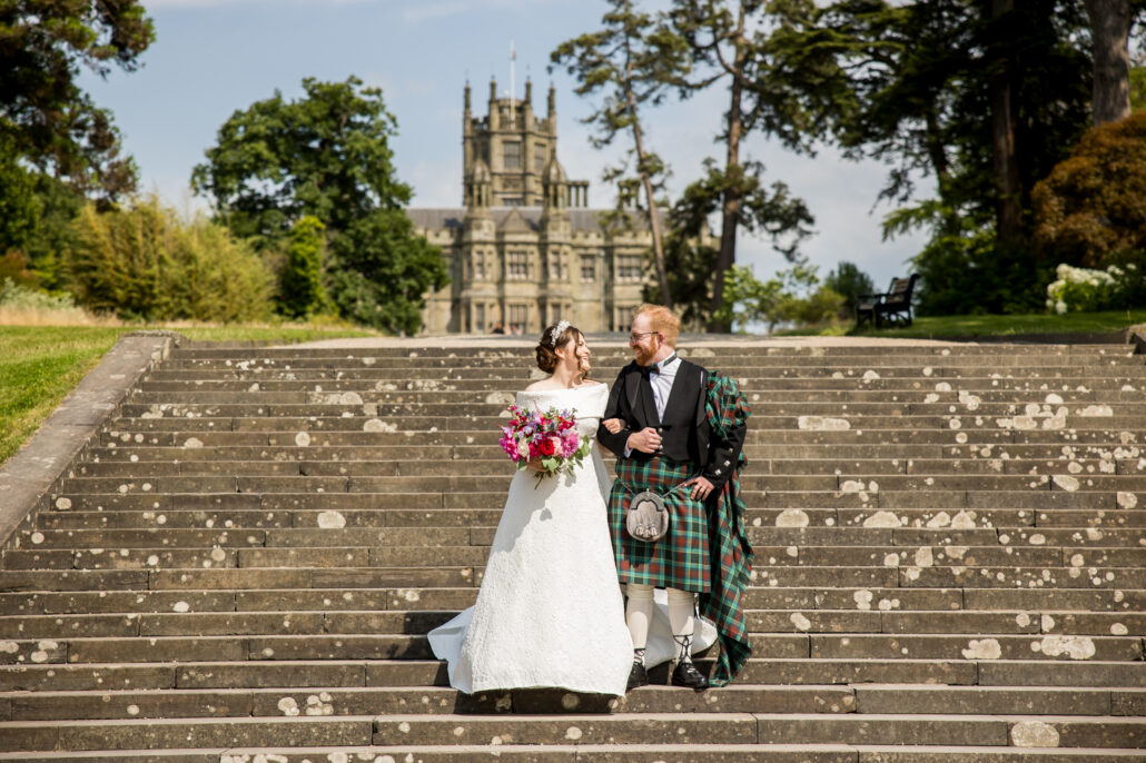 Bride and groom walk down stone steps in formal attire; she holds pink flowers, and he wears a kilt. Captured by a South Wales wedding photographer, a historic building and trees serve as the backdrop.