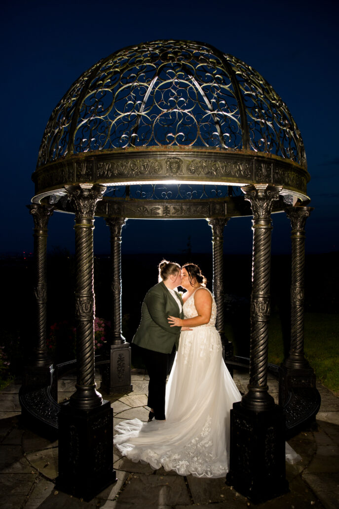 A couple shares a romantic moment under an illuminated wrought iron gazebo at night, surrounded by a stone floor, perfectly captured by a South Wales wedding photographer.