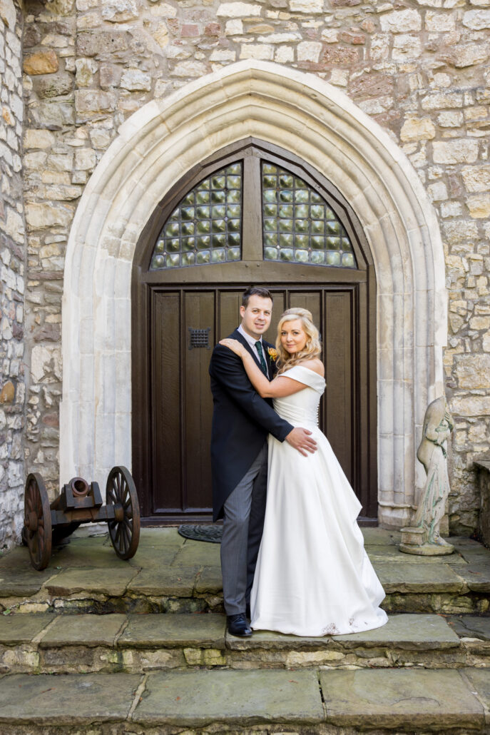 A couple in formal attire stands on stone steps, captured beautifully by a South Wales wedding photographer, in front of an arched door set in a stone building.