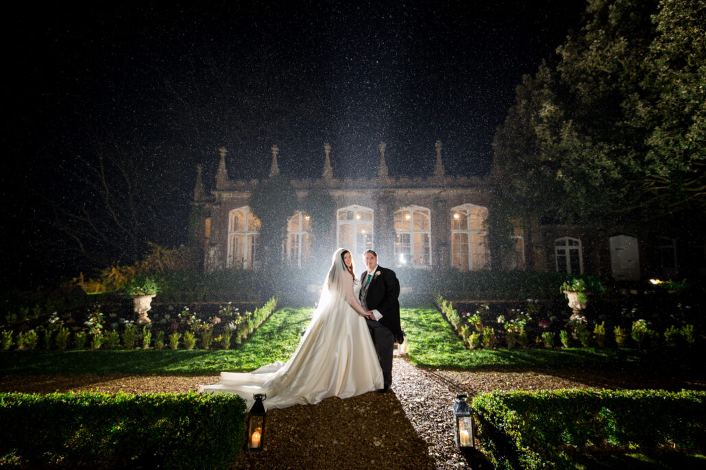 A bride and groom stand before a beautifully illuminated estate in South Wales at night, with a garden path and lanterns leading up to them, perfectly captured by their wedding photographer.