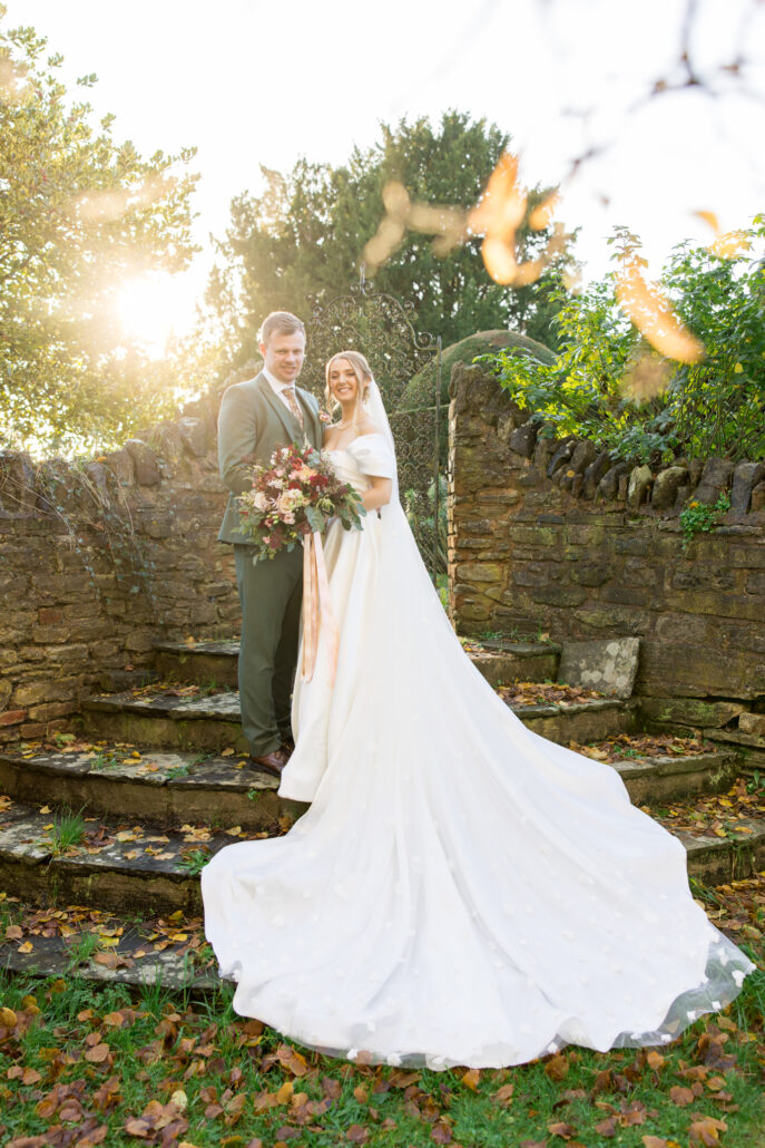 A bride and groom stand on stone steps outdoors, captured beautifully by a South Wales wedding photographer. The bride holds a bouquet and wears a long white gown with a veil, as sunlight shines through the surrounding trees.