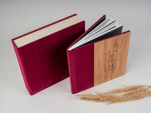Two photo albums with red spines; one closed, the other open with visible pages. The open album has a wooden cover with the date "26 09 24" printed on it. Dry grass lies nearby.