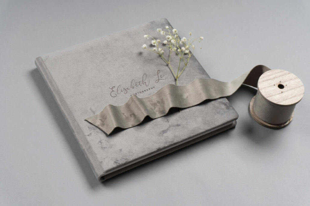 A gray photo album with "Elizabeth L" on the cover, decorated with a sprig of white flowers. A spool of gray ribbon lies next to it.