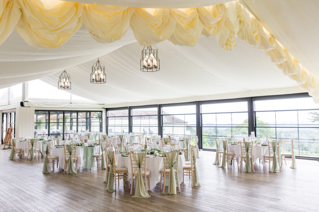 A bright, elegant wedding reception hall with round tables set with green and white decor. Large windows and draped fabric ceiling, with hanging lantern lights.