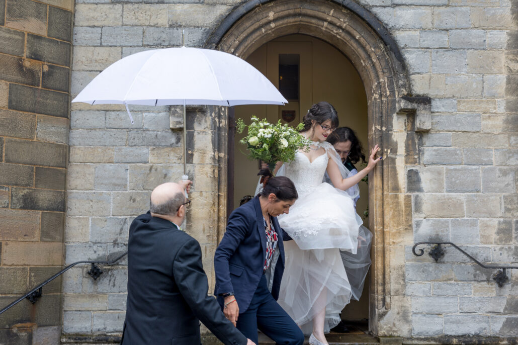 A bride steps out of a stone building, assisted by two people. A man holds a white umbrella over her as she descends the stairs, carrying a bouquet of white flowers.