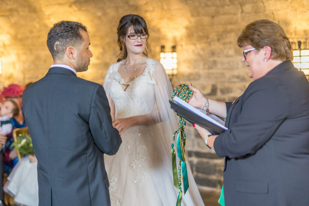 A bride and groom stand before an officiant holding a book and ribbons during a wedding ceremony in a stone-walled room.