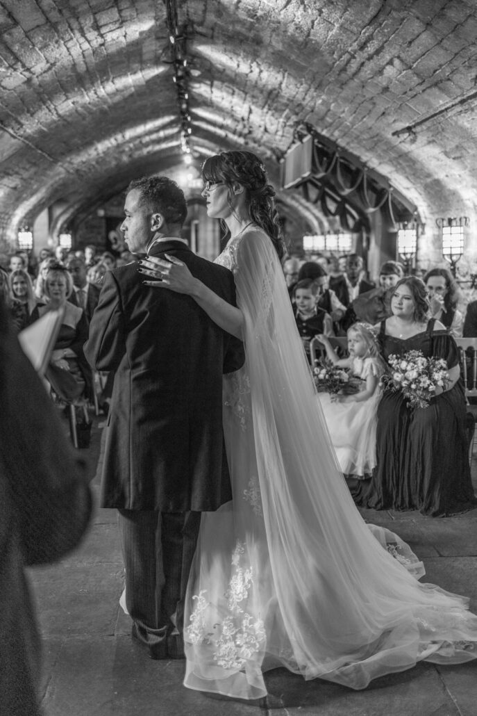 A bride and groom stand together at a wedding ceremony in a dimly lit stone hall with seated guests watching.